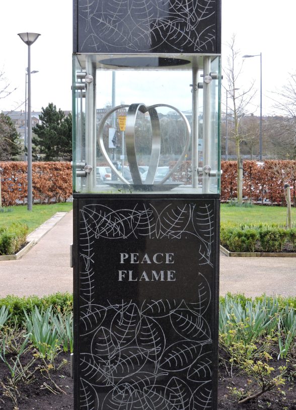 Glass enclosed sculpture on black pillar that says the words Peace Flame