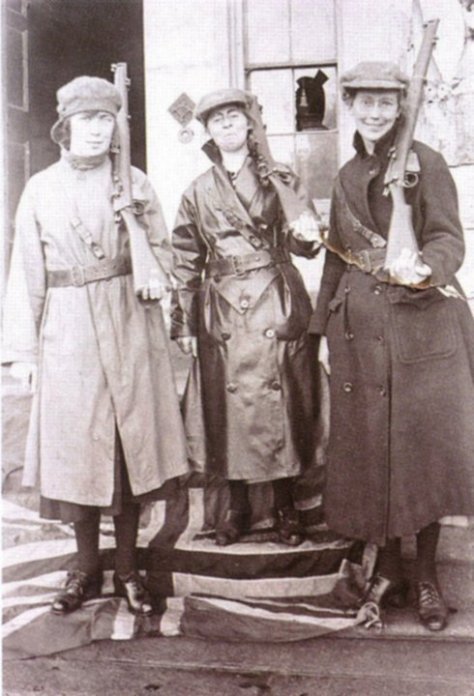 A grainy, sepia-toned photo taken in 1921 of 3 women holding rifles on their shoulders while standing on a British flag.